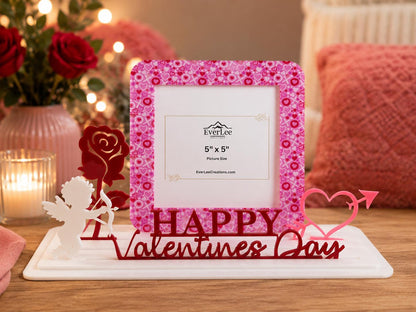 A square picture frame with a pink heart pattern, featuring a white cupid figure, a red rose, and a pink heart with an arrow. The base displays the red text "HAPPY VALENTINES DAY". The frame insert indicates a 5" x 5" picture size.